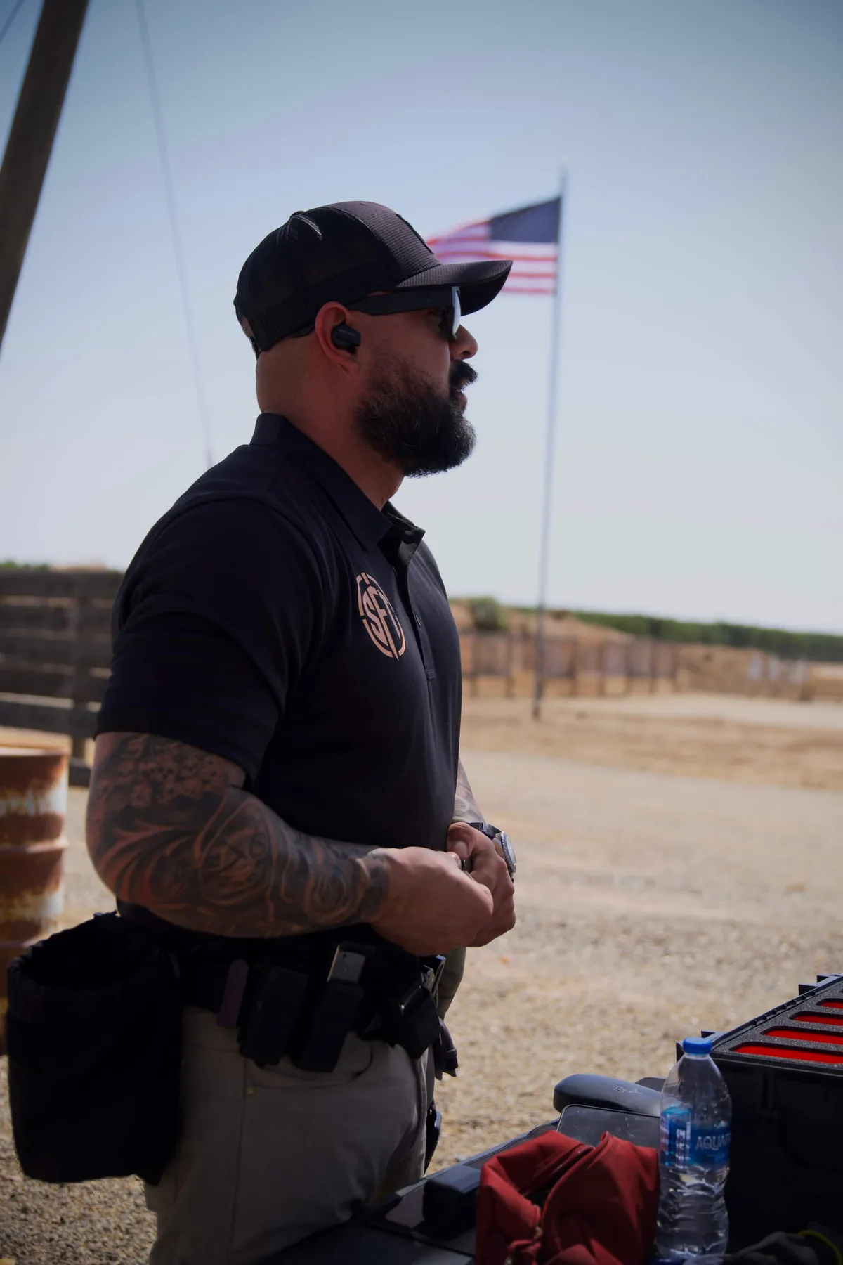 Silverline firearms instructor on the line at Exeter Gunslingers, duty belt and magazines rigged, American flag flying behind the berm.