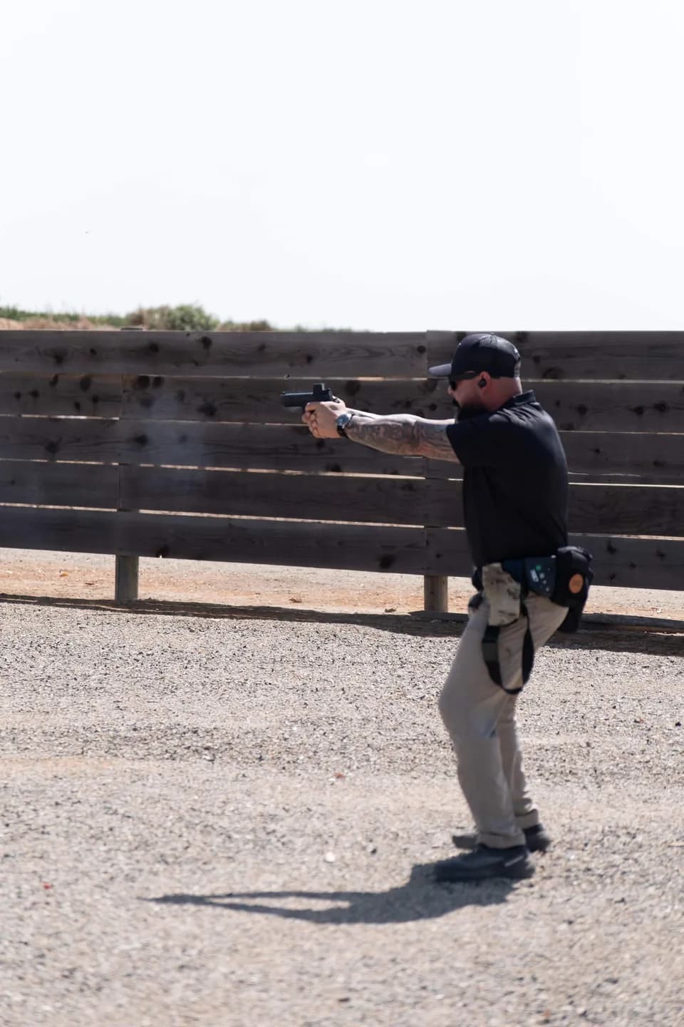 A shooter in a cap and duty belt firing a handgun from the line toward the timber backstop, his shadow stretched across the gravel.