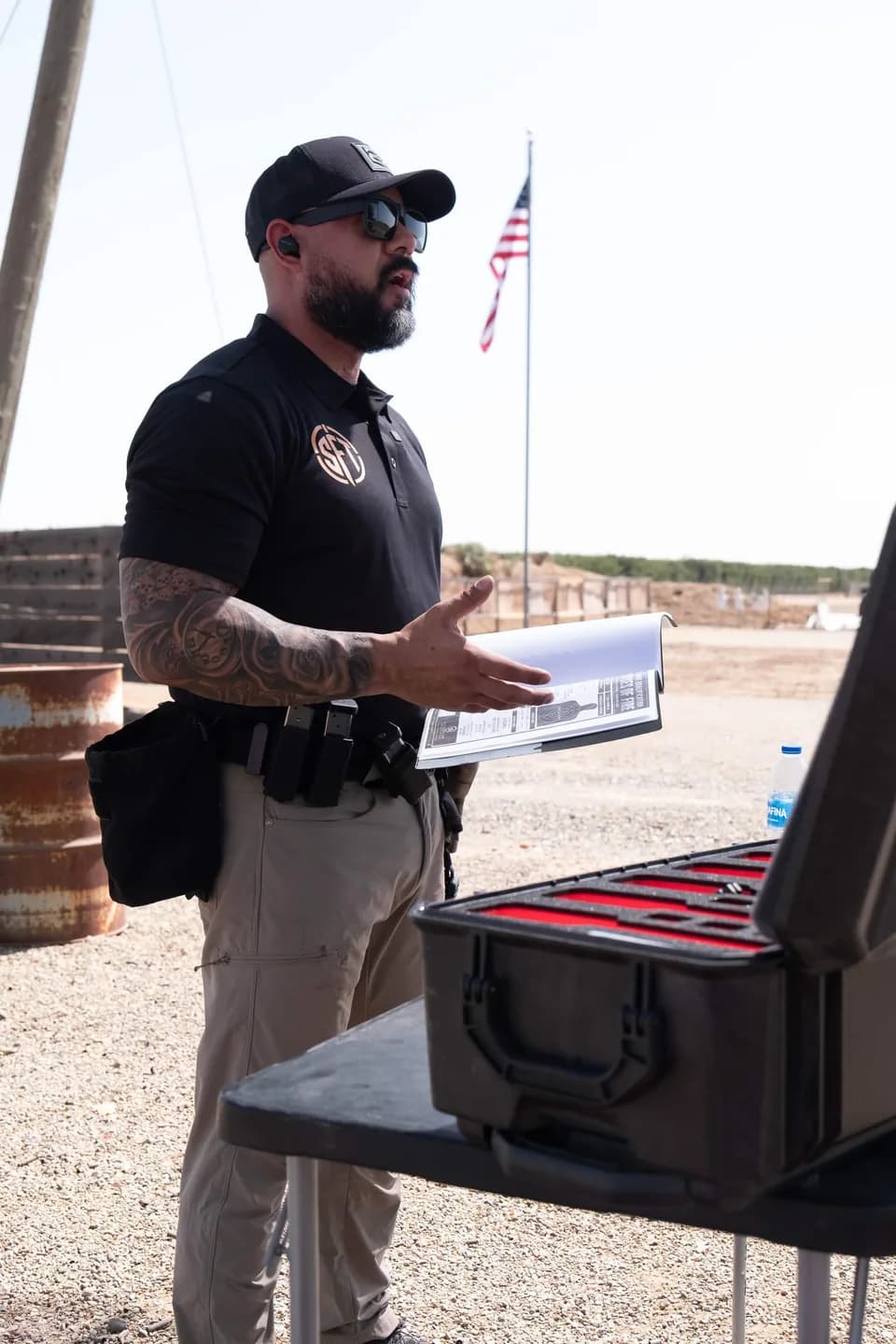 Silverline instructor at the range table with a binder open in one hand, explaining with the other, an open pistol case on the table and the flag behind him.